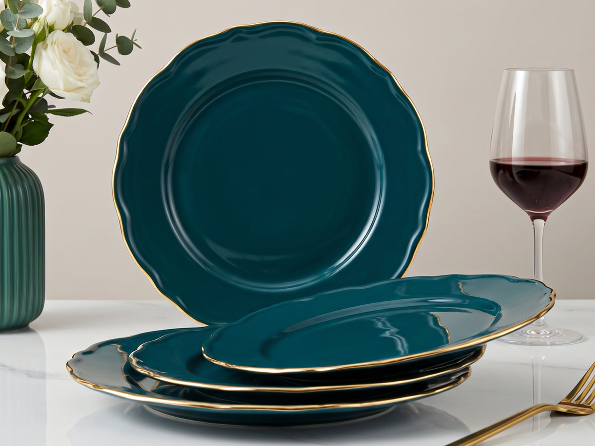 A set of deep teal dinnerware with gold trim, including a large round platter standing upright and several matching plates stacked in the foreground, styled on a white table with a wine glass, flowers, and a fork.