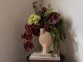 A sculptural beige ceramic vase with a rounded organic shape, styled with a rich floral arrangement of burgundy orchids, red anthuriums, green ferns, and pale yellow hydrangeas, resting on stacked books against a neutral wall.