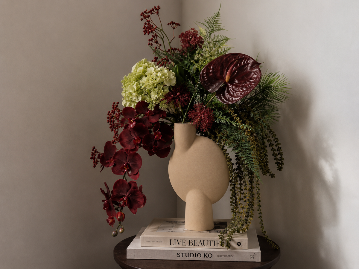 A sculptural beige ceramic vase with a rounded organic shape, styled with a rich floral arrangement of burgundy orchids, red anthuriums, green ferns, and pale yellow hydrangeas, resting on stacked books against a neutral wall.